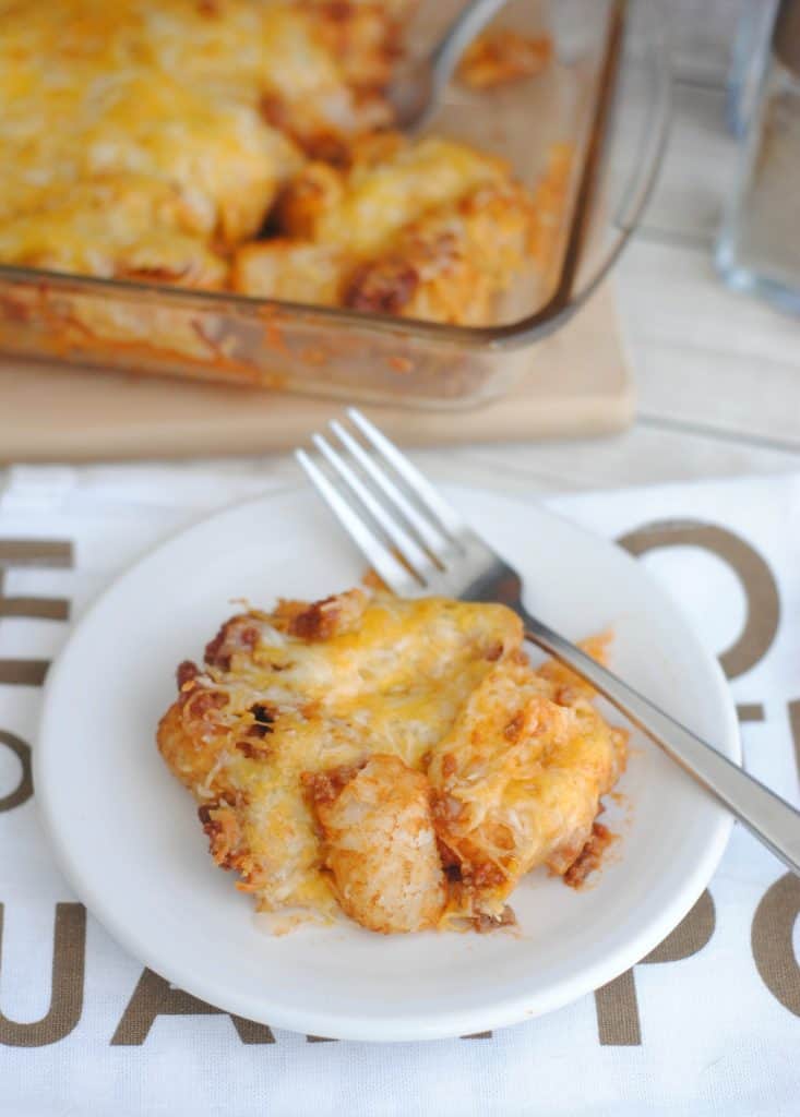 Sloppy Joe Tater Tot casserole on a white plate with a fork with the baking dish in the background. 