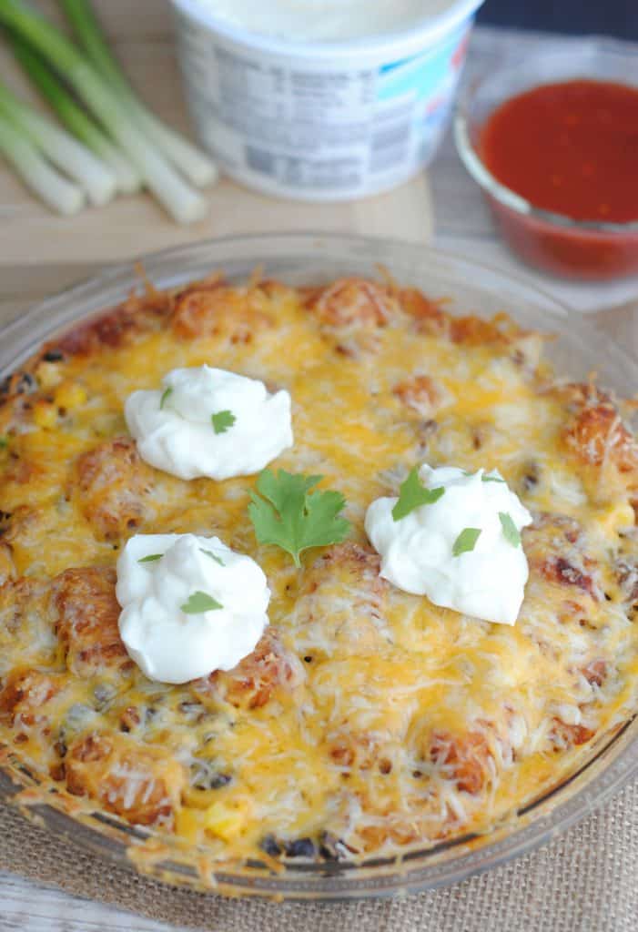 Taco tater tot casserole in a pie pan next to sour cream and green onions