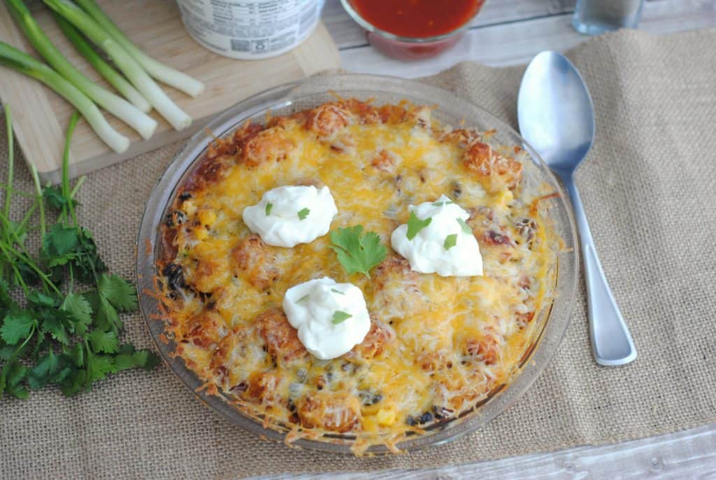 tater tot taco casserole in a pie dish on a burlap mat with a spoon, green onions, and sour cream. 