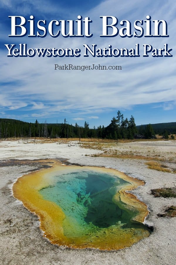 Orange and green thermal pool along Biscuit Basin boardwalk in Yellowstone National Park, blue skies and trees in the background. 