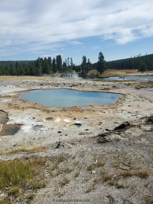 Murky blue thermal pool with brown rocks and thermal areas surrounding it along Biscuit Basin boardwalk, blue skies and trees in the background. 