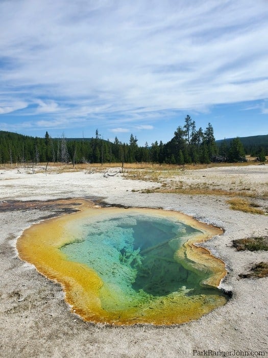 bright orange and green thermal pool with green trees in the background and blue skies with clouds