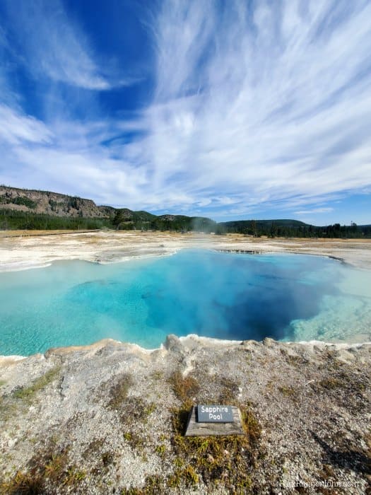 Bright blue almost turquoise Sapphire Pool with clouds over it and green trees in the background along Biscuit Basin boardwalk 