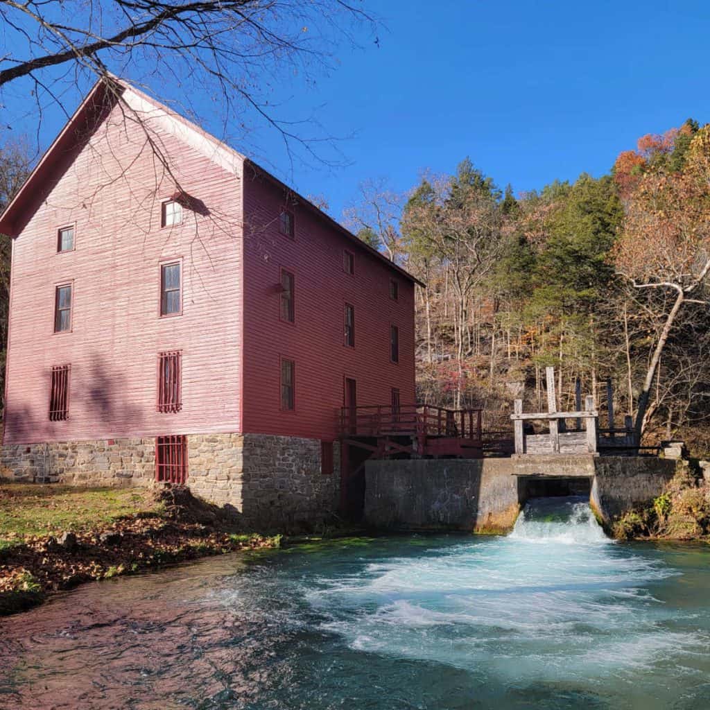 red painted Alley Spring and Mill at Ozark National Scenic Riverway with water coming out of the mill, surrounded by trees and blue skies