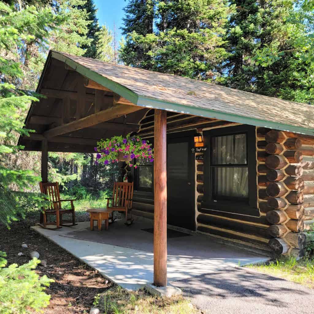 Cabin at Jenny Lake Lodge with a front roof covered front porch with flower baskets hanging down with 2 wooden rocking chairs and table.