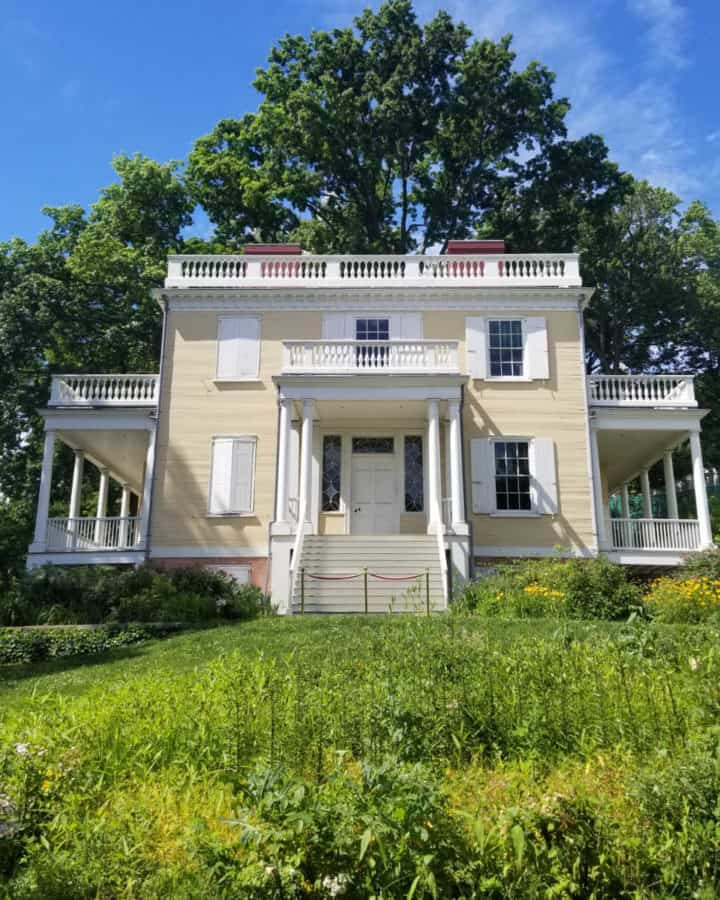 Historic two story house with steps leading up to it on a grassy hill with a tree behind it