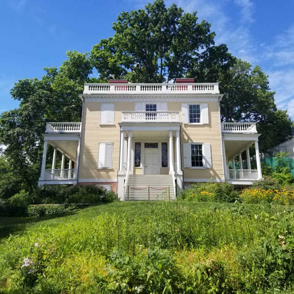 Historic two story house with steps leading up to it on a grassy hill with a tree behind it