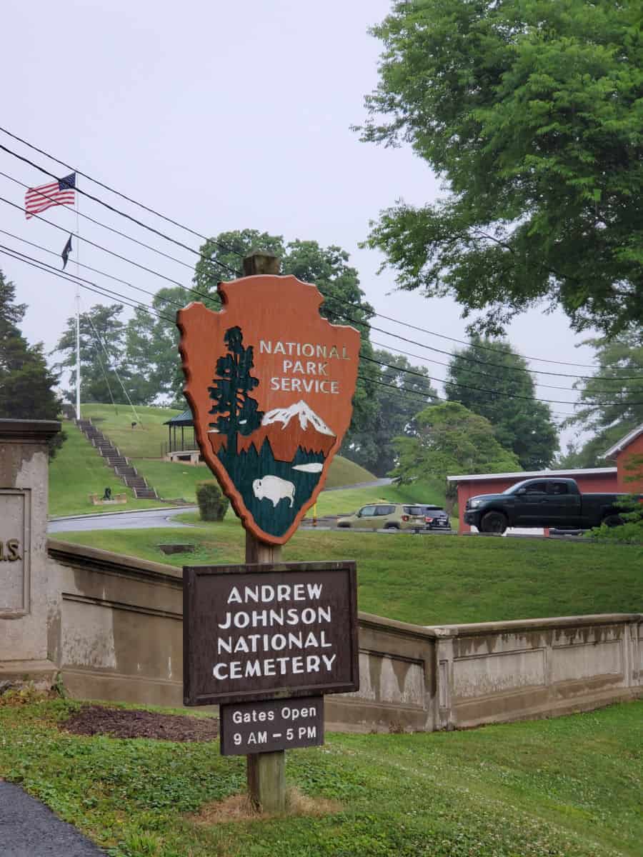 National park service emblem over a sign for the Andrew Johnson National Cemetery with cars and grass in the background