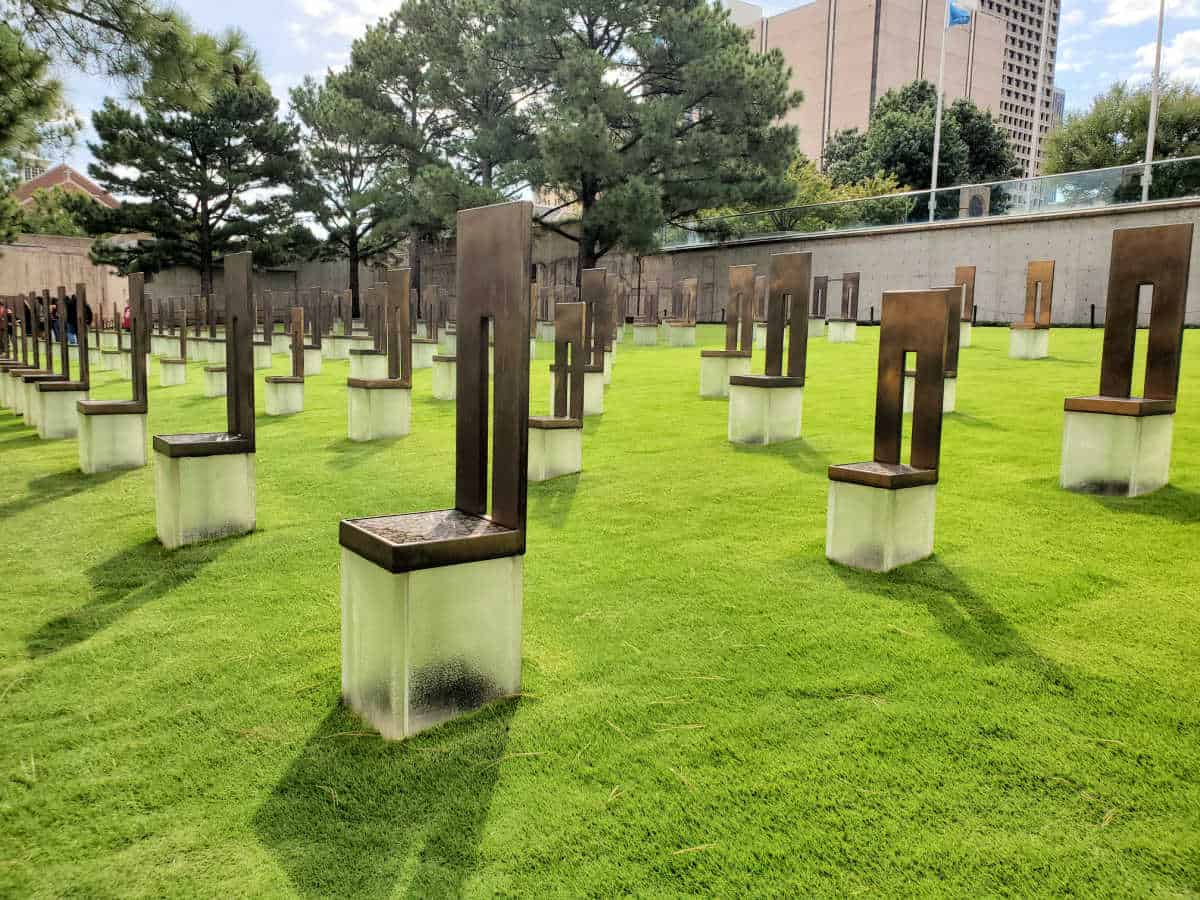 empty chair sculptures in a grass field with buildings in the background