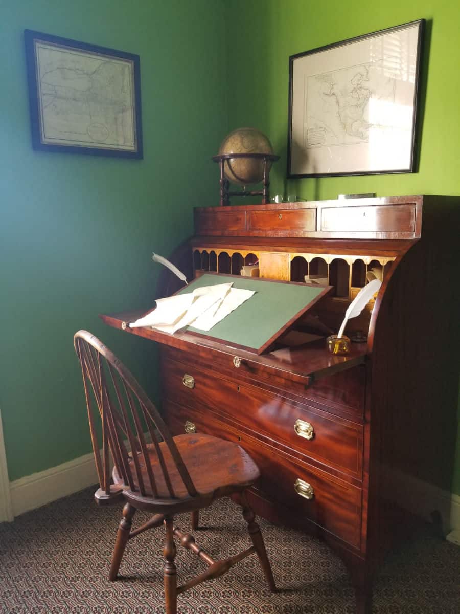Historic wood desk with papers on it, a globe and feather pens, next to a green wall with a wood chair