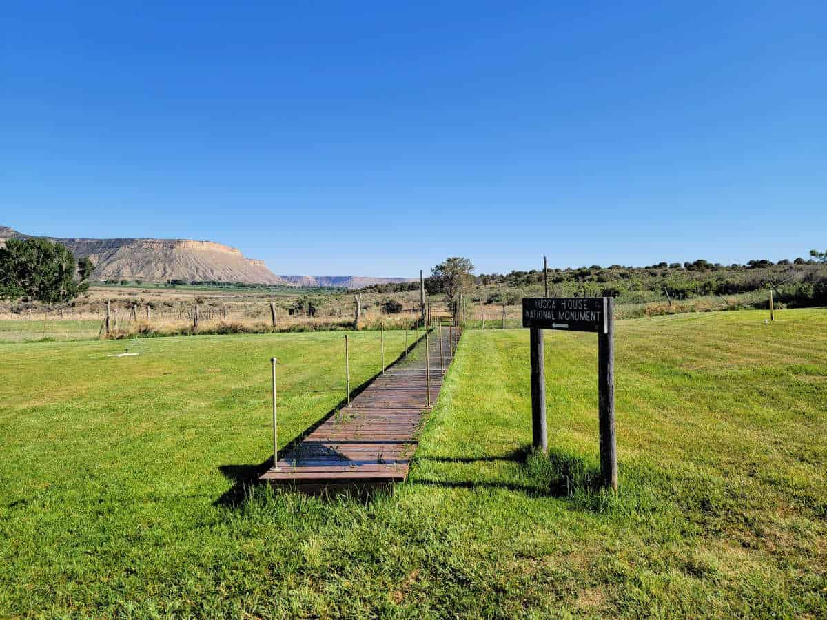 Wooden path with a sign for Yucca House National Monument surrounded by green grass