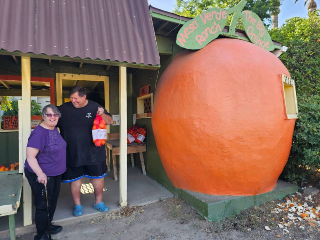 john holding a bag of oranges next to his mom and a large orange statue at a fruit stand