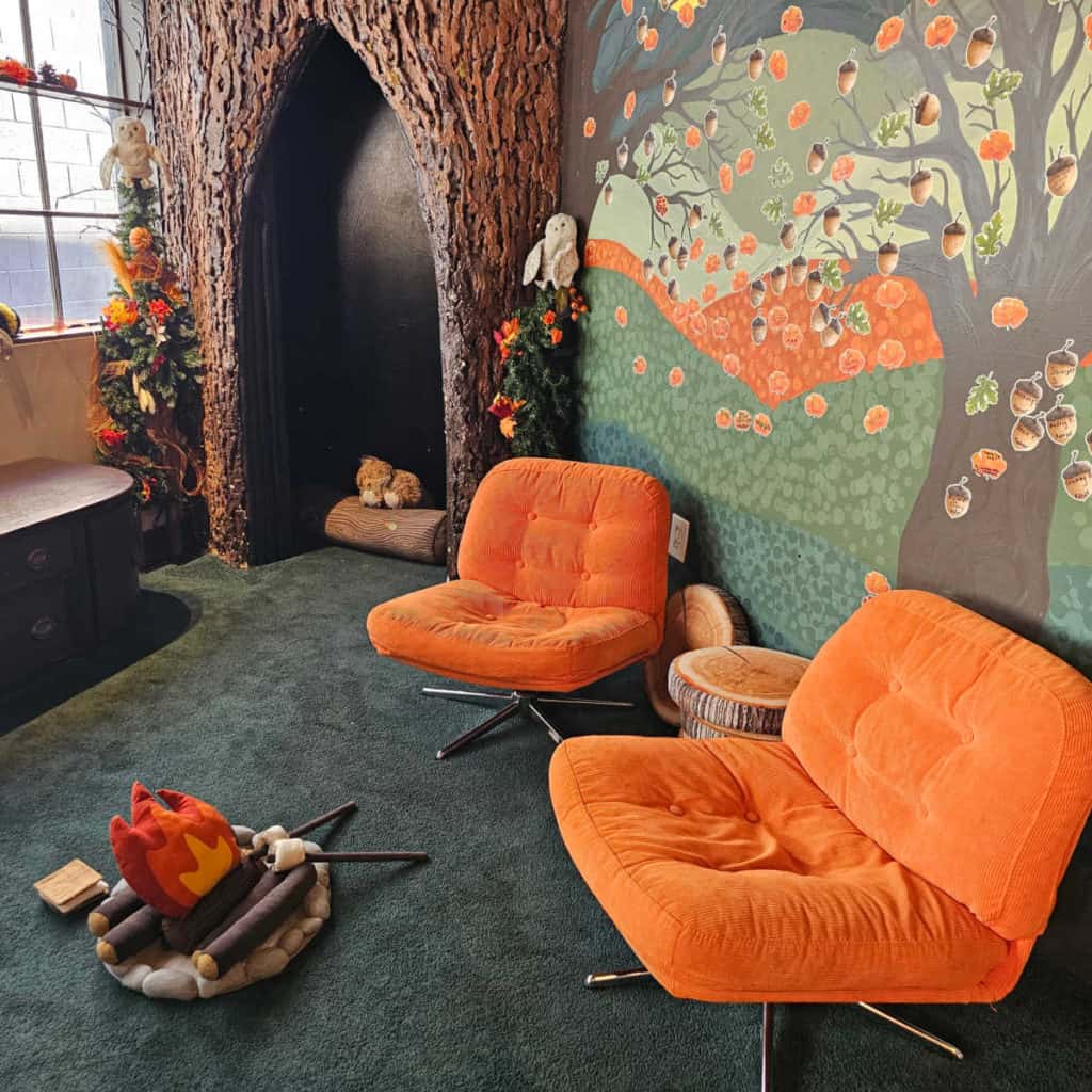 woodsy themed kids play area with orange chairs, a plush fire and nook in a tree with a log pillow