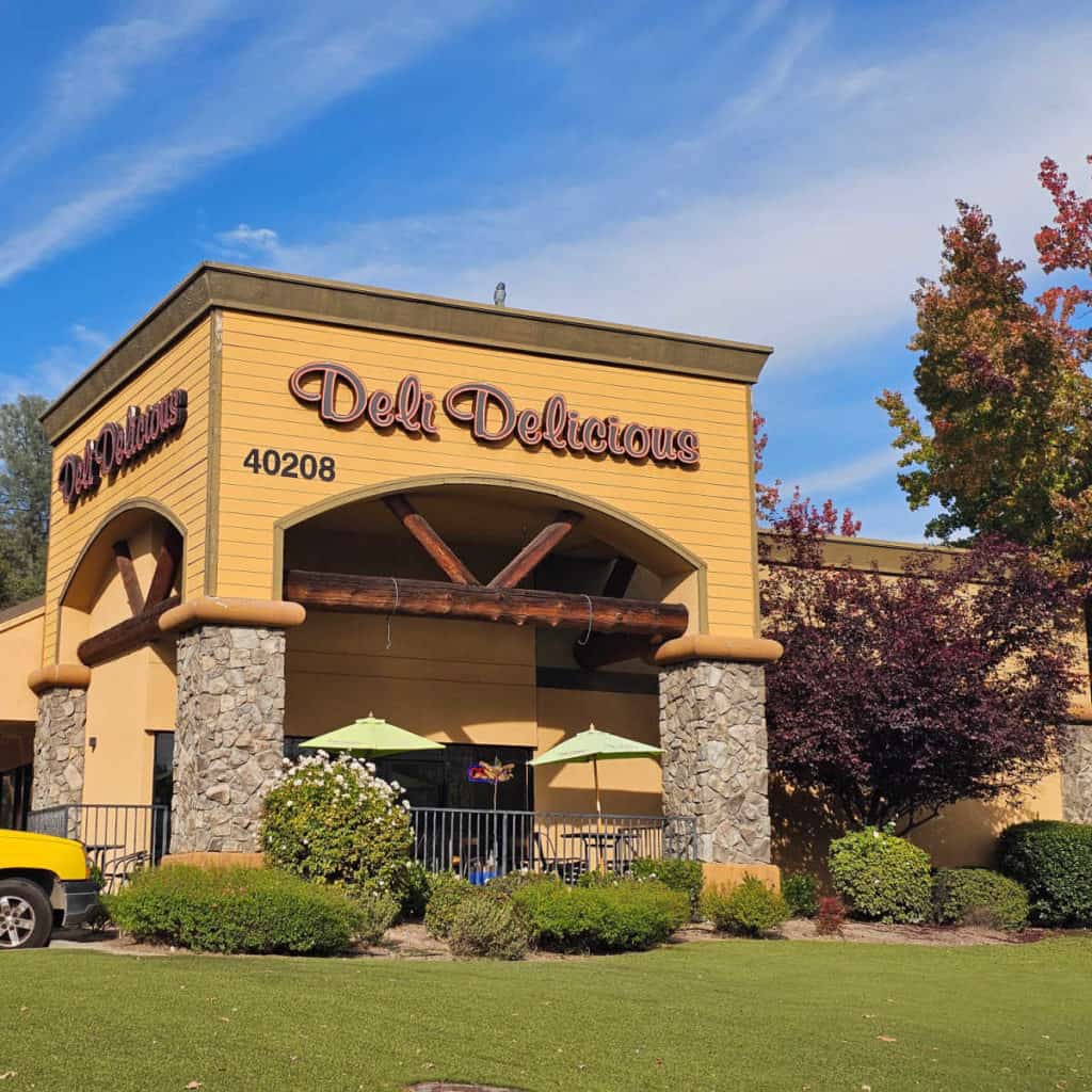 Deli Delicious restaurant exterior with sign, bushes and trees near the outdoor seating